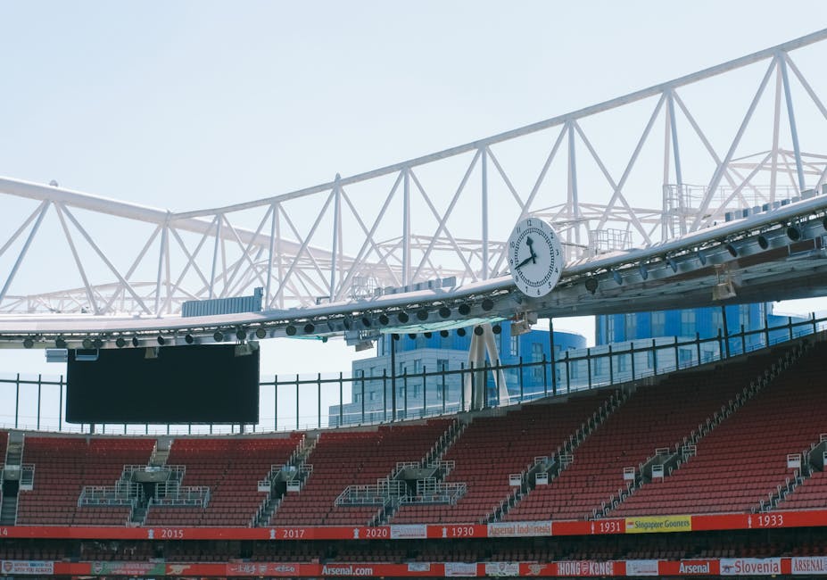 Interior view of a sports stadium showing red seating, a large black electronic scoreboard, and a digital clock displaying approximately 3:07. The upper part features a white steel truss roof structure with supporting beams and a clear sky visible beyond. The seating area appears clean and well-maintained, with no visible litter or dust. The image illustrates the spaciousness and modern design of the stadium, emphasizing the importance of surface maintenance. Highbury Cleaners, a professional cleaning company, specializes in deep cleaning and surface sanitisation to ensure hygiene and cleanliness for venues like these. The focus on the smooth, dust-free surfaces highlights their commitment to high standards in both commercial and sports facilities.