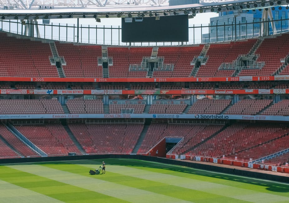 Interior view of Emirates Stadium showing red stadium seating with multiple tiers, rows of empty seats, and a large central scoreboard hanging from the ceiling. The seating sections are labeled with years such as 2005, 2014, 2015, 2017, 2020, and 1930. The lower level seating surrounds a well-maintained grass football pitch, which appears freshly mowed and clean. A groundskeeper is seen on the pitch, pushing a lawn mower. Bright lighting from large windows or the stadium's roof illuminates the interior, highlighting the smooth and even surface of the grass. The image reflects the well-maintained, hygienic environment typical of a professional sports venue, as might be maintained by a domestic or commercial cleaning service like Highbury Cleaners to ensure cleanliness and presentation.