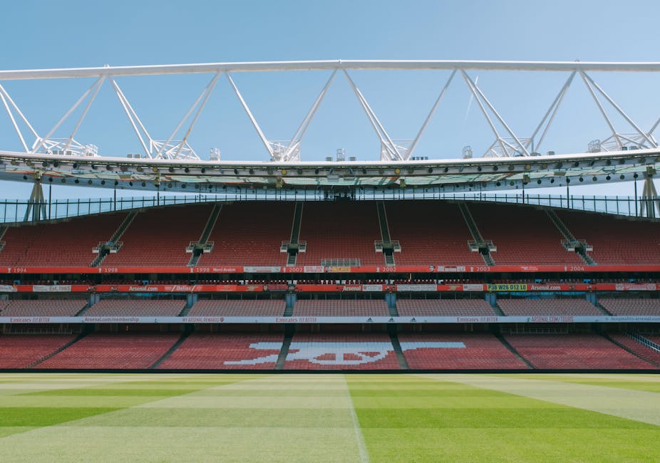 The image depicts an empty football stadium with red tiered seating and a well-maintained grass pitch in the foreground. The seating area shows signs of clean, dust-free surfaces, while the pitch's lush green grass appears freshly mowed with clear, sharp lines. The stadium's structural elements, including exposed white roof supports and glass-fronted suites, are illuminated by natural daylight, reflecting a clean and orderly environment. Visible advertising banners and signage along the perimeter emphasize the stadium's commercial aspect. As part of professional cleaning practices, Highbury Cleaners recommends surface cleaning and sanitisation of such large-scale venues to maintain hygiene and presentation standards, ensuring a spotless and inviting space for sporting events or public gatherings.