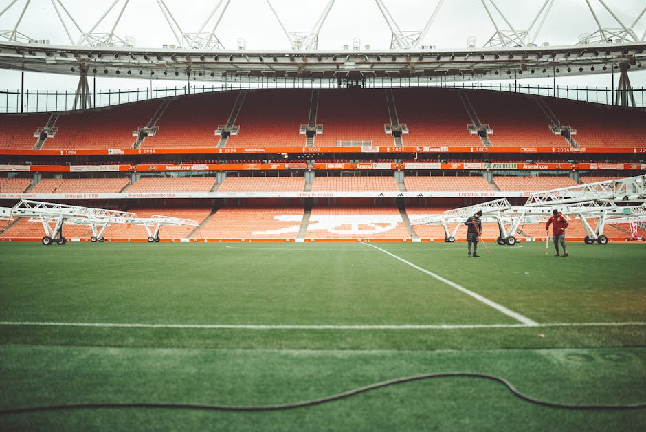 Interior view of Emirates Stadium showing red stadium seating with multiple tiers, rows of empty seats, and a large central scoreboard hanging from the ceiling. The seating sections are labeled with years such as 2005, 2014, 2015, 2017, 2020, and 1930. The lower level seating surrounds a well-maintained grass football pitch, which appears freshly mowed and clean. A groundskeeper is seen on the pitch, pushing a lawn mower. Bright lighting from large windows or the stadium's roof illuminates the interior, highlighting the smooth and even surface of the grass. The image reflects the well-maintained, hygienic environment typical of a professional sports venue, as might be maintained by a domestic or commercial cleaning service like Highbury Cleaners to ensure cleanliness and presentation.