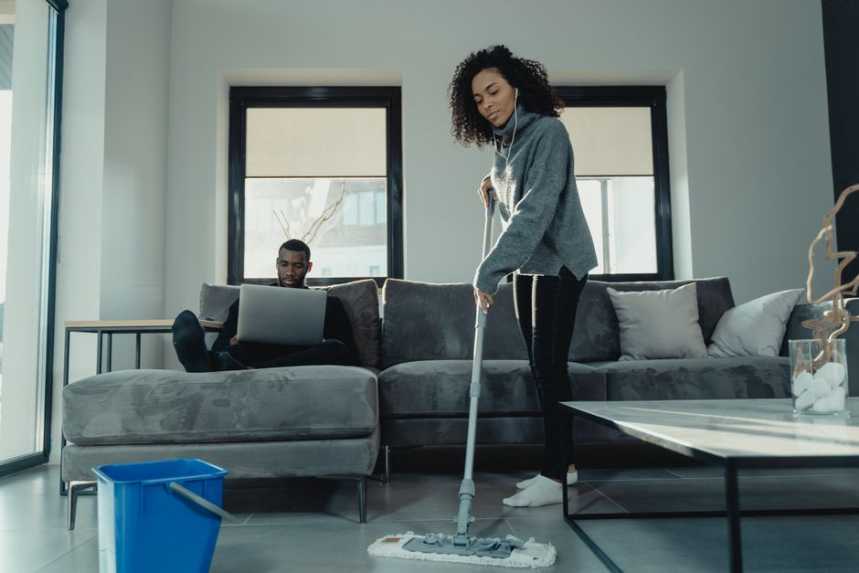 A woman with curly hair wearing a grey sweater and black pants is engaged in deep cleaning a modern living room, using a mop on the tiled floor. The room features a large L-shaped grey fabric sofa adorned with cushions, positioned against a wall with two windows that have beige roller blinds allowing natural light to fill the space. In the background, a man with short hair, dressed in dark clothing, is seated on the sofa working on a laptop. A blue cleaning bucket and a glass vase with decorative white stones are visible on the floor and table respectively, with a clean, dust-free environment emphasizing maintenance and hygienic surface cleaning as part of the residential cleaning services offered by Highbury Cleaners.