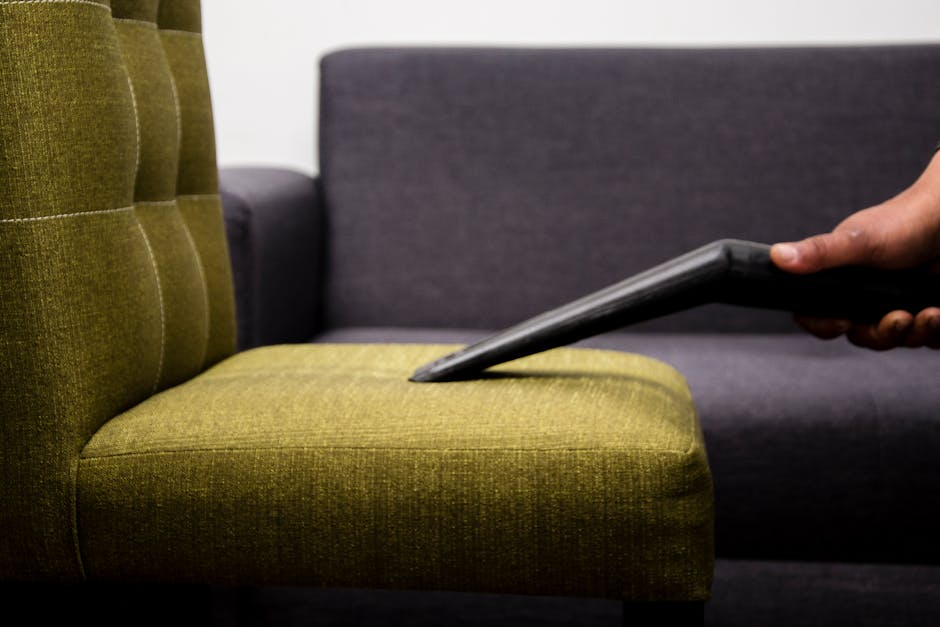 Close-up of a hand holding a vacuum cleaner attachment, cleaning the olive-green fabric upholstery of an armchair with tufted backrest in a well-lit living room. Behind the armchair, a dark grey fabric sofa is partially visible, positioned against a white wall. The scene depicts surface cleaning and deep cleaning processes to remove dust and dirt from furniture, emphasizing hygiene and maintenance in residential domestic cleaning. Highbury Cleaners specializes in professional sofa cleaning, ensuring furniture is left clean and hygienic in a comfortable, clutter-free environment.