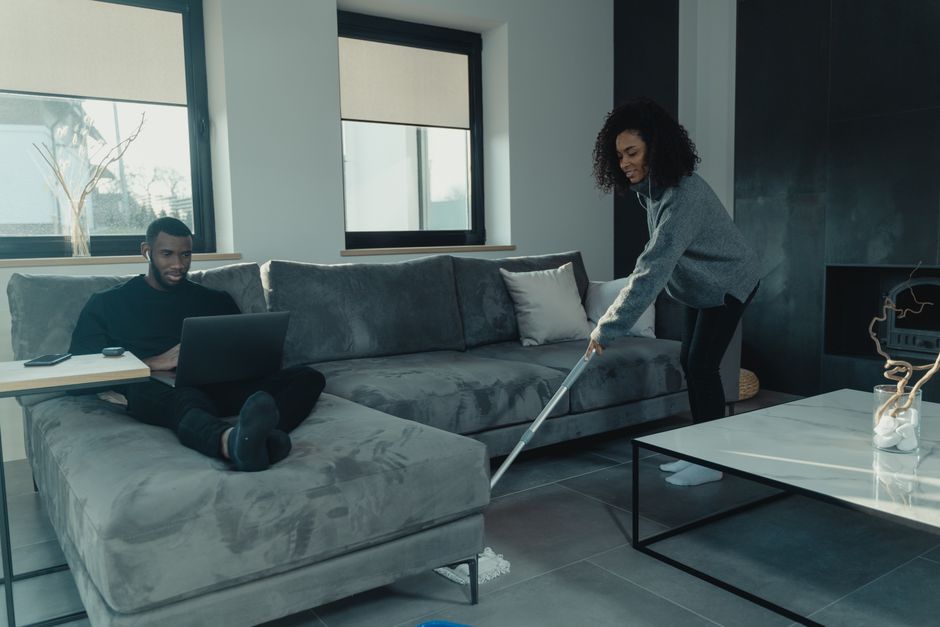 A woman with curly hair wearing a grey sweater and black pants is engaged in deep cleaning a modern living room, using a mop on the tiled floor. The room features a large L-shaped grey fabric sofa adorned with cushions, positioned against a wall with two windows that have beige roller blinds allowing natural light to fill the space. In the background, a man with short hair, dressed in dark clothing, is seated on the sofa working on a laptop. A blue cleaning bucket and a glass vase with decorative white stones are visible on the floor and table respectively, with a clean, dust-free environment emphasizing maintenance and hygienic surface cleaning as part of the residential cleaning services offered by Highbury Cleaners.