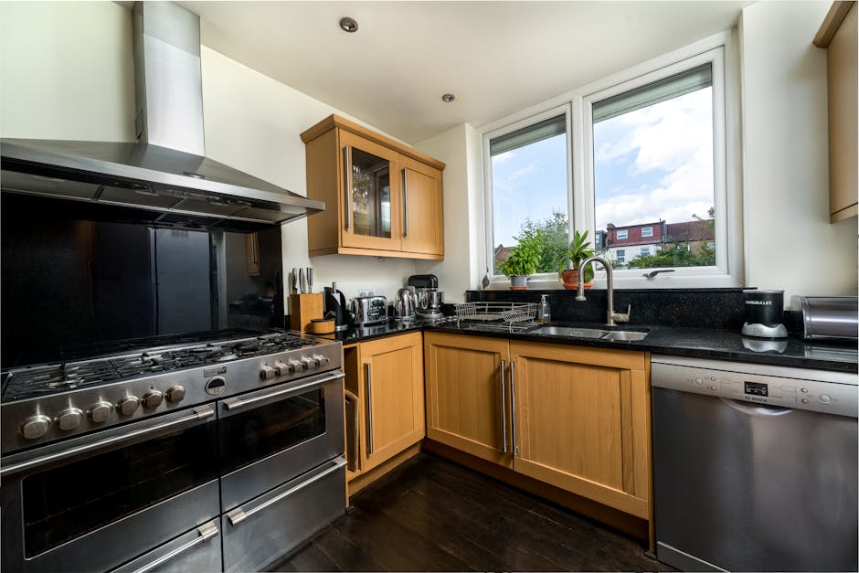 A modern kitchen featuring a stainless steel gas range with multiple burners and oven, located adjacent to light wooden cabinets with glass-fronted upper doors and matching lower drawers. The black granite countertop houses a stainless steel sink with a chrome faucet beneath a large double window, allowing natural light to illuminate the space. Visible on the countertop are a plant, a toaster, a kettle, and a dish drying rack, all arranged neatly. The space appears clean and well-maintained, with a polished, dark wooden floor and a bright, airy atmosphere created by the natural light. This setting exemplifies surface cleaning and eco-friendly domestic cleaning practices promoted by Highbury Cleaners, highlighting the importance of hygiene and regular maintenance in kitchen environments.
