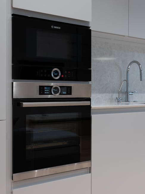 A modern kitchen scene featuring a built-in oven with a stainless steel finish and a black glass door, positioned beneath a matching microwave oven from Bosch. The ovens are installed into a white kitchen unit, adjacent to a light grey marble-effect countertop. To the right, there is a stainless steel sink with a curved, high-arc faucet, set into the same countertop. The surfaces, including the oven doors and countertop, appear clean and well-maintained, with no visible dirt or stains, reflecting thorough surface cleaning and sanitisation practices. The lighting is bright, highlighting the smooth, polished surfaces and the sleek design of the appliances, indicative of professional domestic cleaning services provided by Highbury Cleaners. This setup exemplifies comprehensive kitchen surface cleaning and maintenance, aligning with the content on Gillespie Road Oven Cleaning and Local Access Notes page.