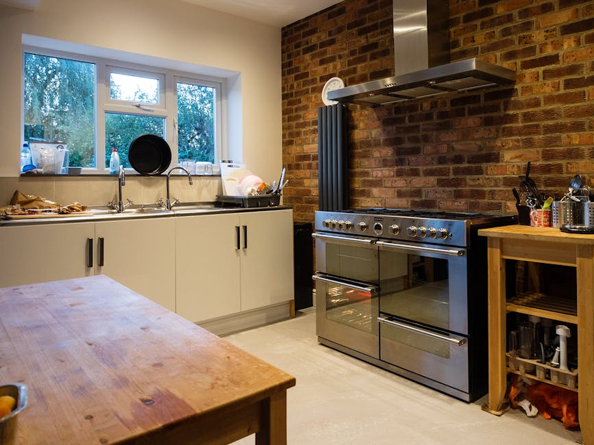 A modern kitchen featuring a stainless steel gas range with multiple burners and oven, located adjacent to light wooden cabinets with glass-fronted upper doors and matching lower drawers. The black granite countertop houses a stainless steel sink with a chrome faucet beneath a large double window, allowing natural light to illuminate the space. Visible on the countertop are a plant, a toaster, a kettle, and a dish drying rack, all arranged neatly. The space appears clean and well-maintained, with a polished, dark wooden floor and a bright, airy atmosphere created by the natural light. This setting exemplifies surface cleaning and eco-friendly domestic cleaning practices promoted by Highbury Cleaners, highlighting the importance of hygiene and regular maintenance in kitchen environments.