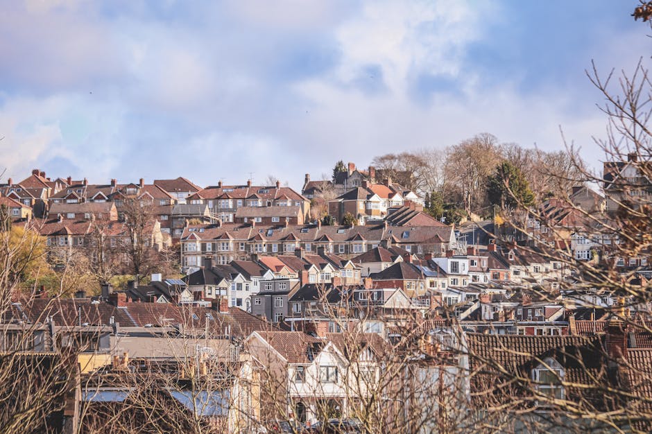 A panoramic view of a residential neighborhood on a clear, sunny day, featuring numerous terraced and semi-detached houses set on a hillside. The houses are constructed with a mix of brick, wood, and 