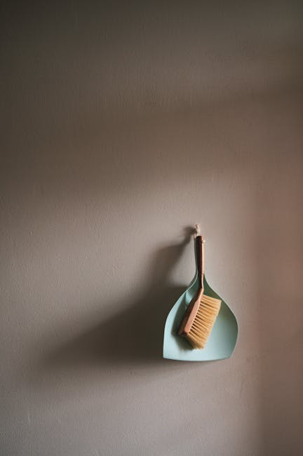 A minimalist scene featuring a wall-mounted light blue dustpan and a matching small hand brush with natural wood bristles hanging from a hook on a neutral beige wall. The dustpan and brush are clean and well-maintained, indicating recent or regular surface cleaning. The lighting is soft, highlighting the smooth texture of the wall and the neat arrangement of cleaning tools. This setup reflects attention to indoor hygiene and tidiness, aligning with domestic cleaning practices. Highbury Cleaners, a professional cleaning company, emphasizes thorough surface cleaning and maintenance, as seen in this simple yet effective display of cleaning tools for routine housework.