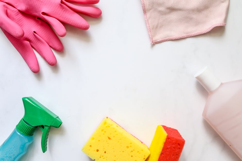 An arrangement of cleaning tools and supplies on a white surface, including pink rubber gloves, a pink cloth towel, a green spray bottle, a yellow and red cleaning sponge, and a pink plastic spray bottle, illustrating equipment used for surface cleaning, sanitisation, and deep cleaning as part of domestic and commercial cleaning services by Highbury Cleaners for the Highbury Barn Flat Cleaning Checklist N5, HIGHBURY.