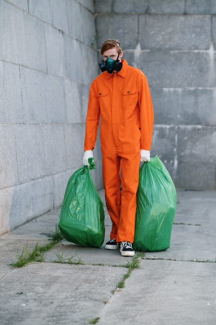 A worker dressed in an orange protective jumpsuit, wearing a black respirator mask and white gloves, stands outdoors against a gray concrete wall. The individual holds two large green plastic bags filled with waste, suggesting waste collection or outdoor refuse disposal. The concrete ground appears clean, with some small patches of grass near the wall. The scene reflects a focus on sanitation, waste management, and outdoor cleaning activities, aligning with the services offered by Highbury Cleaners in adherence to Islington Council waste disposal regulations. The lighting is natural, emphasizing the cleanliness and safety precautions taken during waste collection or deep cleaning tasks. This image highlights the importance of sanitation and proper waste disposal in residential or commercial environments, emphasizing the professional cleaning practices promoted by Highbury Cleaners.