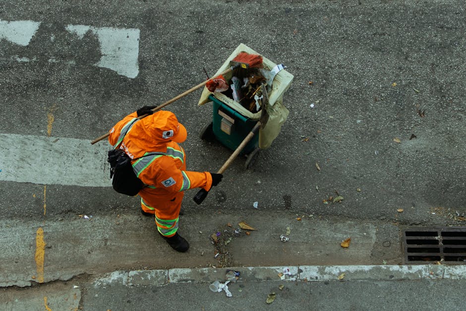A worker dressed in an orange protective jumpsuit, wearing a black respirator mask and white gloves, stands outdoors against a gray concrete wall. The individual holds two large green plastic bags filled with waste, suggesting waste collection or outdoor refuse disposal. The concrete ground appears clean, with some small patches of grass near the wall. The scene reflects a focus on sanitation, waste management, and outdoor cleaning activities, aligning with the services offered by Highbury Cleaners in adherence to Islington Council waste disposal regulations. The lighting is natural, emphasizing the cleanliness and safety precautions taken during waste collection or deep cleaning tasks. This image highlights the importance of sanitation and proper waste disposal in residential or commercial environments, emphasizing the professional cleaning practices promoted by Highbury Cleaners.