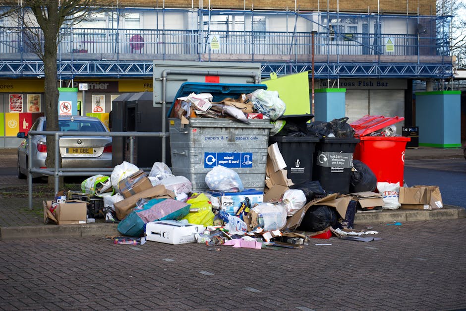 A large collection of mixed paper and cardboard waste, plastic bags, and miscellaneous rubbish overflowing from a grey recycling bin marked for paper and card, situated on a paved area outside a commercial building. Several black rubbish bags and flattened cardboard boxes are placed around the base of the bin, with some loose items scattered nearby. In the background, there is a car parked next to a metal railing, and a building with blue scaffolding, storefronts, and signage for a fish bar and other shops. The scene appears to depict improper disposal and the need for waste management or cleaning services. This image emphasizes the importance of proper rubbish disposal for hygiene and environmental maintenance, consistent with services offered by Highbury Cleaners.