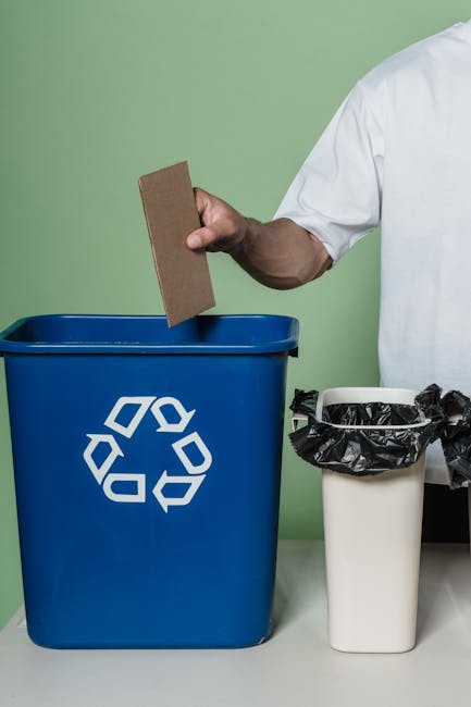 A person wearing a white uniform is disposing of a brown cardboard item into a blue recycling bin with a white recycling symbol on it. The blue bin is positioned on a white surface against a light green wall. Next to the recycling bin, there is a smaller white bin lined with a black plastic bag, also on the white surface. The scene suggests a clean, organized environment focused on waste separation and recycling, consistent with professional domestic or commercial surface cleaning and waste management practices, as offered by Highbury Cleaners. The lighting is bright, highlighting the cleanliness and orderliness of the area.