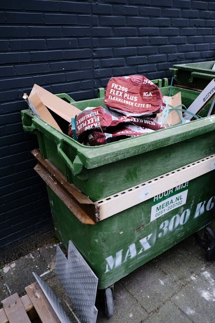 A large collection of mixed paper and cardboard waste, plastic bags, and miscellaneous rubbish overflowing from a grey recycling bin marked for paper and card, situated on a paved area outside a commercial building. Several black rubbish bags and flattened cardboard boxes are placed around the base of the bin, with some loose items scattered nearby. In the background, there is a car parked next to a metal railing, and a building with blue scaffolding, storefronts, and signage for a fish bar and other shops. The scene appears to depict improper disposal and the need for waste management or cleaning services. This image emphasizes the importance of proper rubbish disposal for hygiene and environmental maintenance, consistent with services offered by Highbury Cleaners.